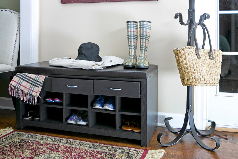 A dark wood entryway shoe storage bench with drawers and cubbies, topped with boots, a hat, and a coat, placed beside a metal coat rack holding a woven handbag in a bright foyer.