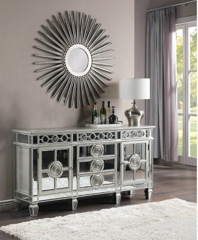 Elegant mirrored sideboard with ornate circular detailing and reflective panels, paired with a modern silver table lamp and decorative bottles, set beneath a striking sunburst mirror in a stylish living room with soft curtains and a plush white rug.