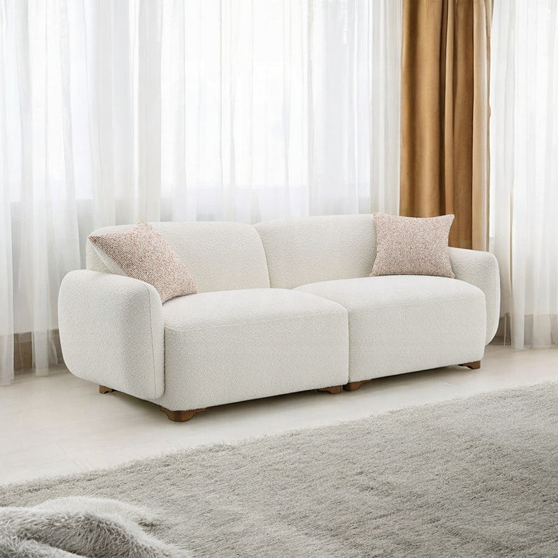 Contemporary white bouclé sofa with two textured beige throw pillows, placed in a bright living room with sheer curtains and a soft area rug.